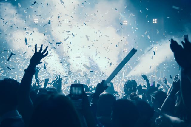 Stock image showing people's silhouette with their arms raised while inside a nightclub with bright sides and confetti falling